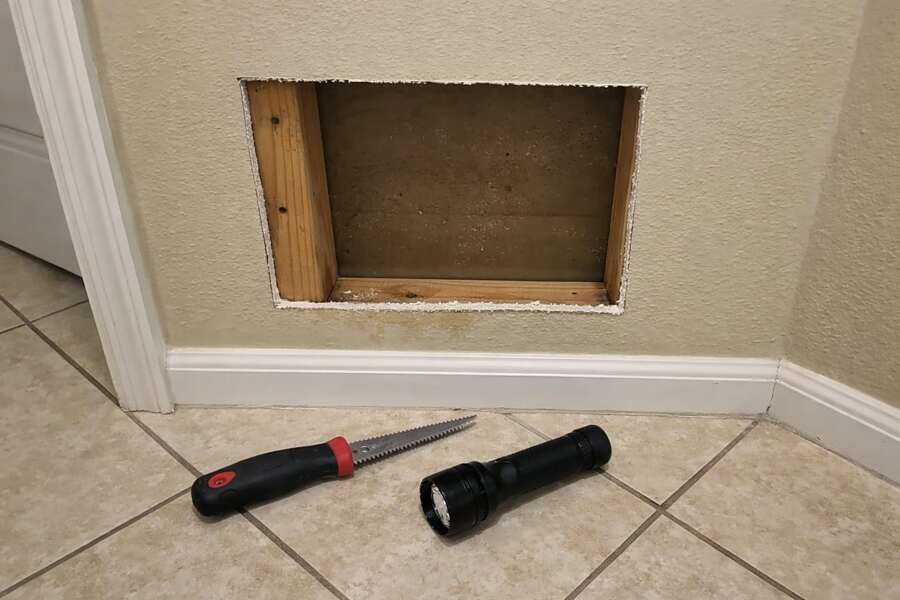 A rectangular access opening cut into hallway drywall with a jab saw and flashlight on the tile below