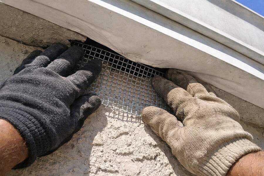 Gloved hands pressing galvanized hardware cloth into a gap between a soffit panel and a stucco wall