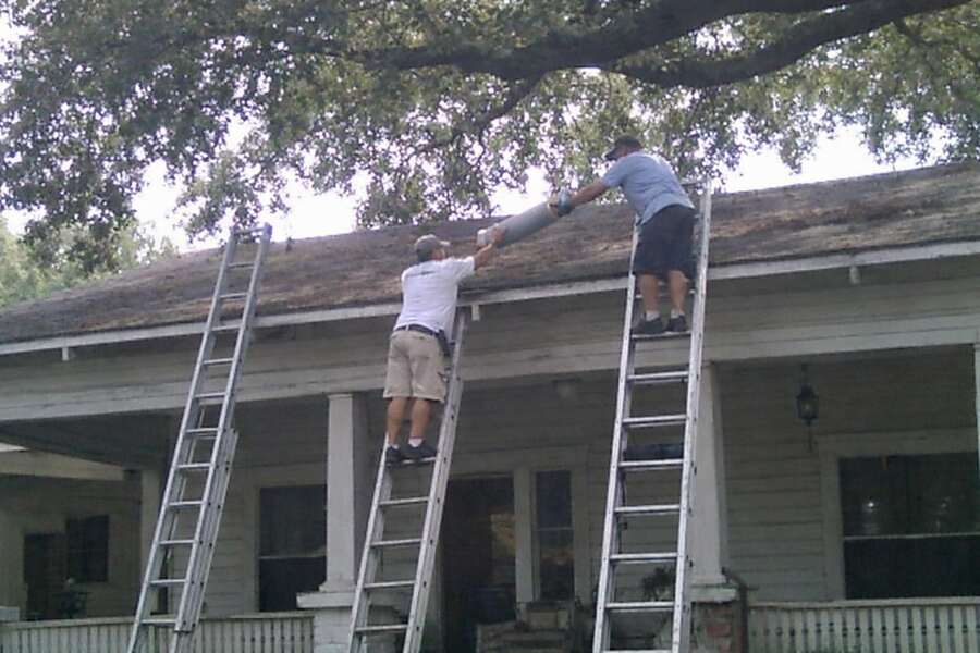 Two roofers on ladders at the roofline of an older wood-frame house with a deep porch
