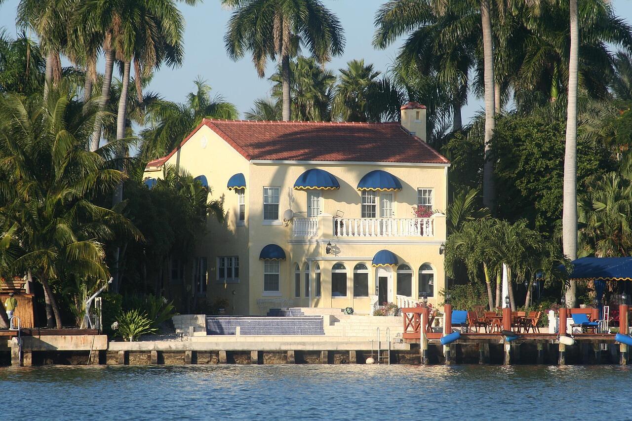 A two-story waterfront house with a tile roof and palms beside a canal